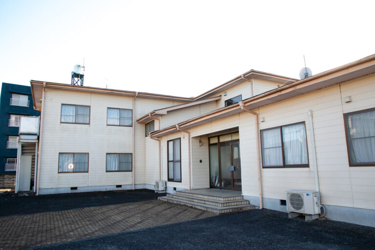 白色サイディングの介護施設建物。駐車場側からの外観で、玄関と複数の窓が見える。